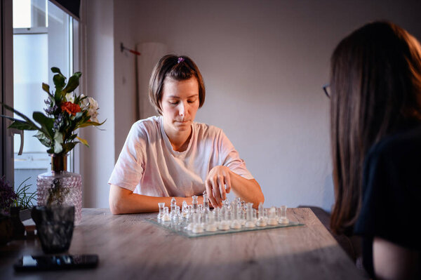 Two individuals play chess with a glass set at a wooden table, surrounded by flowers and natural light from a nearby window.