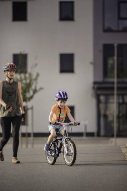 Gülümseyen çocuk kendine güvenen bir bisikletle kaldırımlı bir yolda kaskını ve günlük kıyafetlerini takıp açık havada güneşli bir günün tadını çıkarıyor..
