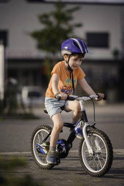 Gülümseyen çocuk kendine güvenen bir bisikletle kaldırımlı bir yolda kaskını ve günlük kıyafetlerini takıp açık havada güneşli bir günün tadını çıkarıyor..