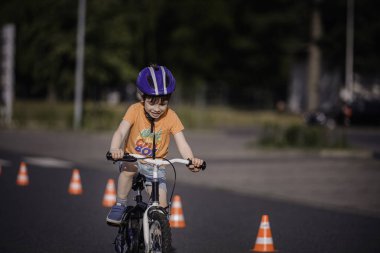 Kask takan genç bir çocuk, bir sıra trafik konisi boyunca küçük bir bisiklet sürüyor, güneşli bir yolda açık hava antrenmanı sırasında odaklanmış ve kararlı..