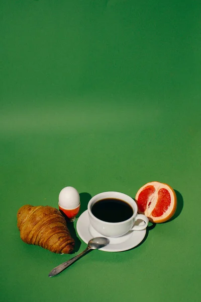cup of coffee and croissant and egg and fruit on a green background