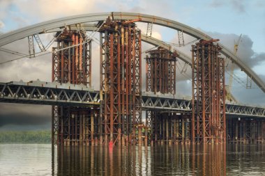 Geniş bir nehir boyunca inşa edilen köprünün arka planında koyu yağmur bulutları olan mavi bir gökyüzü var. 