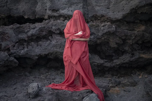 Young woman covered in a long red veil from head to toes looking up with the eyes closed while standing in front of a rocky wall, conceptual eerie apparition, symbolic silence and invisible presence