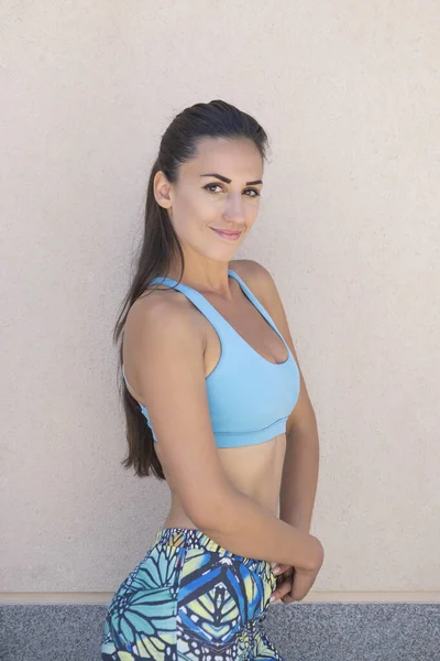 Attractive Caucasian millennial woman wearing sportswear in blue tones and posing after a fitness session looking at the camera and smiling, athletic person showing off her fit and beautiful physique