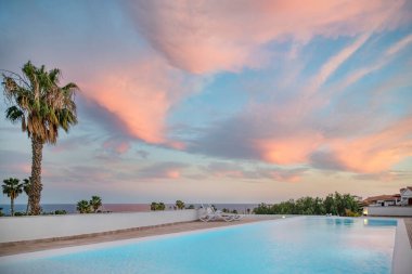 Amarilla Golf, Tenerife, Kanarya Adaları, İspanya - 4 Mart 2023 'te sakin bir tatil köyünün üzerinde alacakaranlık ışığı, etkileyici bir gün batımı gökyüzünü kaplayan dramatik bulutlar ve bir turkuaz yüzme havuzuna yansıyor.