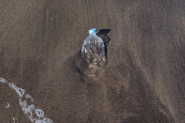Small squashed plastic water bottle washed on the pristine shores with black volcanic sand of the island of Tenerife, marine debris contaminating and polluting the oceans and the coastal environment
