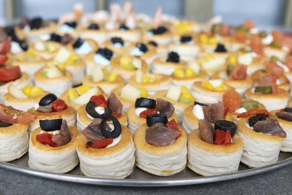 Large serving plate filled with a variety of Mediterranean canapes with focus on savory vol-au-vent pastries filled with anchovies, black olives and red peppers, bite-sized snacks for a party or event