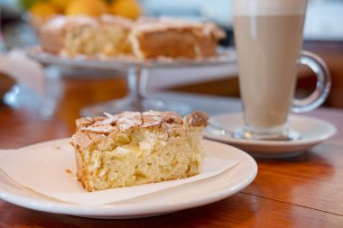 Büyük bir bardak sütlü kahveyle birlikte bir tabakta lezzetli bir dilim kek, üzerinde elma ve badem süngeri olan lezzetli bir tatlı, üzerinde şeker ve fındık gevreği ve büyük bir kahveyle servis ediliyor.