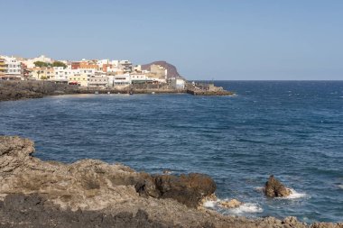 Engebeli volkanik sahil manzarası ve derin mavi Atlantik Okyanusu ve kıyı boyunca uzanan renkli binaları ile büyüleyici balıkçı köyü Los Abrigos, Tenerife, Kanarya Adaları, İspanya