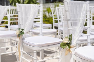 Close-up of an elegant seating arrangement for a wedding or a romantic ceremony featuring white chairs and cushions, draped in sheer fabric and set in place with rose bouquets tied with golden ribbons clipart