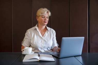 Orta yaşlı bir kadın, elinde bir fincan kahve, açık bir planlamacı ve minimalist çalışma ortamı ile çevrili sakin ve profesyonel davranışlı bir bilgisayarın ekranına bakıyor.