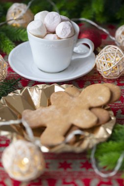 Sıcak çikolatayla dolu beyaz bir fincanın yer aldığı ve üstü yumuşak beyaz ve pembe şekerlemelerle kaplı ve yanında zencefilli kurabiyelerle dolu bir tabakla birlikte kutlanan Noel sahnesi.