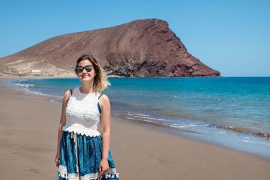 Playa de la Tejita 'nın altın kumlarında duran mutlu genç kadın, Montana Roja' ya tepeden bakan kalabalık olmadan ve güneşli Tenerife, Kanarya Adaları, İspanya