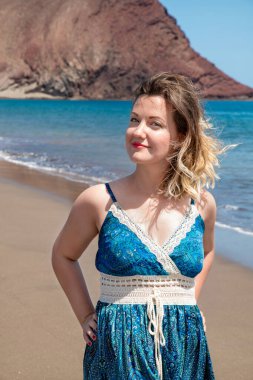 Güzel volkanik plajda duran çarpıcı mavi bir elbisesi olan genç bir kadın. Elleri kalçalarında ve güzel bir gülümsemeyle rahatlamış görünüyor. La Tejita sahilinde, Tenerife, Kanarya Adaları, İspanya.