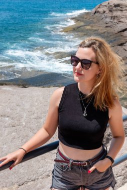 Kolsuz siyah bir bluz giyen genç bir kadın ve burgundy kemerli yüksek atık kot şortlu sakin ve rahat bir ifadeyle kayalık bir kıyı şeridinin yanında durup tatilin tadını çıkartıyor.