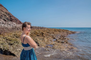 Playa de la Tejita 'nın kıyılarında mavi desenli elbiseler giyen güzel bir kadın. Montana Roja volkanik konisinin dibinde duruyor. Okyanusa bakıyor.