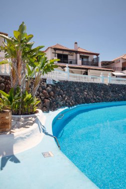 Paraiso Royal, Tenerife, Kanarya Adaları, İspanya - 22 Haziran 2025: tropik bitkiler ve geleneksel Kanarya tarzı tatil daireleriyle çevrili turkuaz-mavi sularla dolu yüzme havuzu