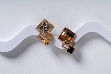 Dalgalı beyaz bir yüzeye yerleştirilmiş karmaşık detaylarla geometrik tasarımlar içeren zarif bir çift altın küpe. Modern mücevher işçiliği ve stili sergileniyor