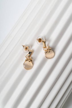 Zarif çiçek desenleri ve dairesel aksanları olan zarif altın küpeler beyaz desenli bir yüzeye yerleştiriliyor. Karmaşık işçilik ve şık bir çekicilik sergiliyor.