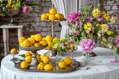 Limonlar süslü tabaklarda, yuvarlak bir masada canlı çiçek aranjmanlarının yanında sergileniyor. Meyve ve çiçeklerin karışımı göz alıcı ve dengeli bir kompozisyon yaratır.
