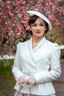 Beyaz takım elbiseli ve şapkalı bir kadın pembe dalların altında zarif bir şekilde poz veriyor. Kırmızı ruj ve inci küpeler takıyor, sakinliğini ve kendine güvenini gösteriyor.