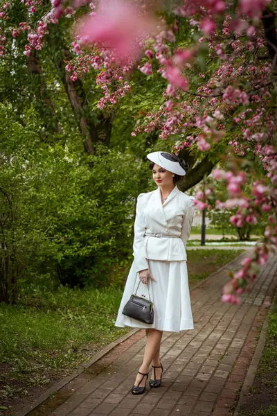 Klasik beyaz kıyafetli, şapkalı zarif bir kadın çiçek açan pembe ağaçların altında zarif ve sofistike bir şekilde duruyor. Elinde, çiçek açan ağaçların altında duran beyaz kıyafetli yemyeşil adamla çevrili bir el çantası var. Yüksek kalite fotoğraf