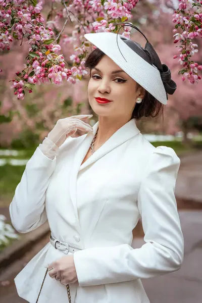 Beyaz takım elbiseli ve şapkalı zarif bir kadın kiraz çiçeklerinin altında özgüvenle poz veriyor. Klasik entellektüellik, sakin bir ifade ve dengeli bir tavır sergiliyor.
