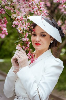 Bahçede çiçek açan pembe çiçeklerle çevrili klasik beyaz kıyafetli zarif bir kadın. Nazikçe bir dal tutuyor, dinginliği ve zarafeti ince bir gülümsemeyle ifade ediyor.