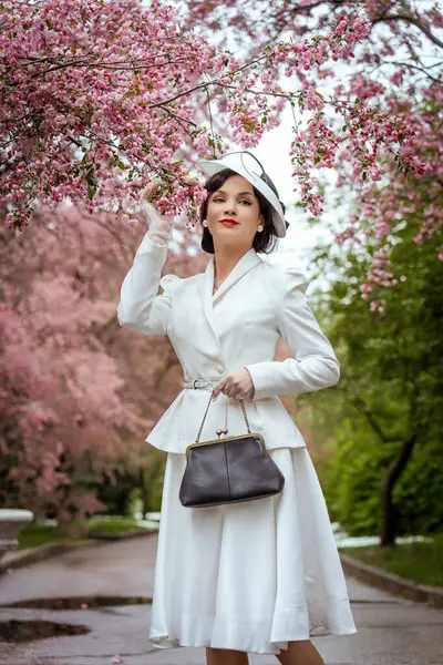 Beyaz elbiseli ve şapkalı bir kadın parkta kiraz ağacının tadını çıkarıyor. Bahar çiçekleri arasında zarafeti ve klasik güzelliği somutlaştıran klasik bir çanta taşıyor.