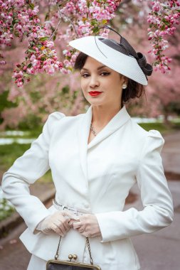 Klasik beyaz takım elbiseli ve şapkalı bir kadın canlı kiraz çiçeklerinin altında zarif bir şekilde duruyor. Antika bir çanta tutuyor ve özgüven ve entelektüellik yayıyor.