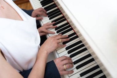 Birlikte piyano çalan bir çiftin yakın çekimleri, anahtarların üzerinde ellerini ve ortak müzik aktiviteleriyle olan bağlantılarını vurgulamak. Her ikisi de sevgi ve birliği simgeleyen yüzükler takıyor.