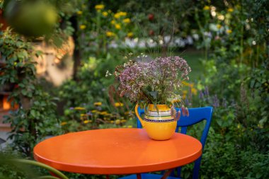 Parlak turuncu masa, sarı bir vazoda çiçek buketi, yemyeşil bir bahçede. Canlı renkler ve doğal ortam canlı ve davetkar bir atmosfer yaratıyor.