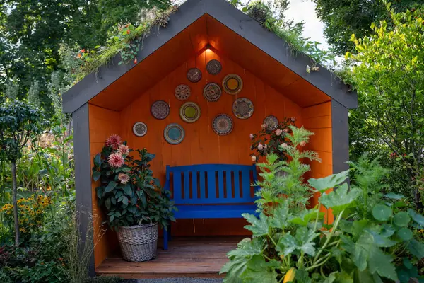Canlı bahçe kulübesi, mavi bir bank, çiçek dekoru ve duvarında dekoratif tabaklar. Yeşilliklerle çevrili bu rahat köşe renkli bir bahçe ortamında huzurlu bir geri çekilme sunuyor.
