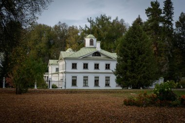 Sonbaharda yemyeşil ormanlarla çevrili tarihi bir konak. Büyük beyaz bina zarif bir şekilde yıkılmış yaprakların ve yükselen ağaçların arasında duruyor, sükunet ve zamansızlık hissi uyandırıyor.