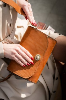 Kırmızı tırnaklı bir kadın şık kahverengi bir deri çanta tutuyor, şık kıyafetler içinde oturuyor. Bu resim moda ve zarafeti vurguluyor, detaylara ve kişisel stile dikkat çekiyor
