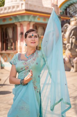 Kadın zarif bir şekilde mavi bir sari içinde poz veriyor, açık havada sakin bir ifadeyle büyütülmüş. Dekor kültürel mimariyi harmanlıyor, canlı geleneksel kıyafetleri geliştiriyor.