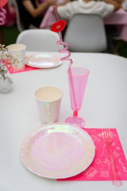 A vibrant table setting features a pink plastic glass with a playful straw, disposable plates, and cups on a white table. Perfect for celebrations with a fun, youthful vibe