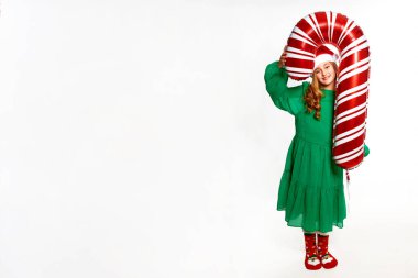 Noel kızı elinde kocaman bir balon tutuyor, yeşil elbise giyiyor, Noel Baba şapkası ve yeni yıl partisi için kırmızı çoraplar giyiyor.