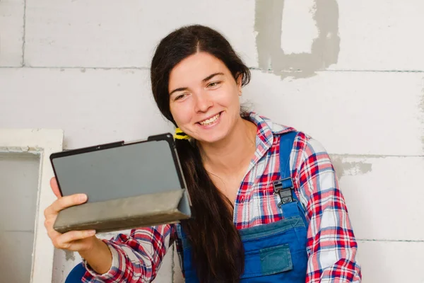smiling woman makes repairs in new apartment by herself. communicates on tablet on wall background