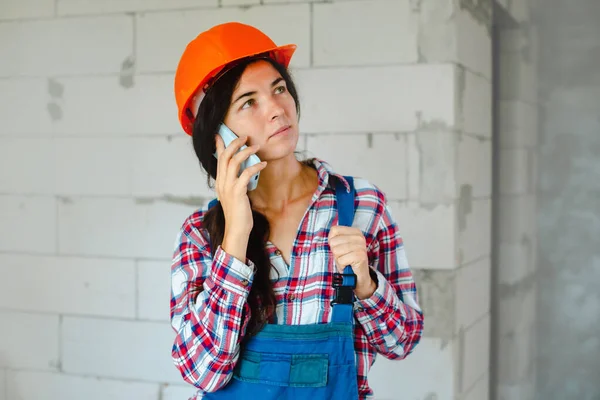 concentrated worker woman in blue overall having phone conversation during break from work at construction site indoors