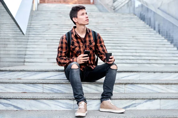 stylish guy with mobile phone and coffee sitting on granite stair. spring or summer trip and walking in city