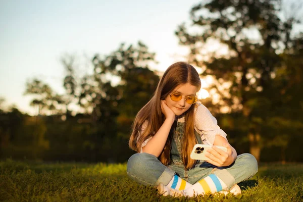 Adorable child girl play and watch video with cell phone sitting in deep grass in park
