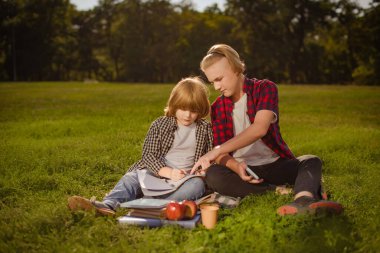 İki çocuk yeşil çimlerde ödev yapıyor. Büyük kardeş küçük olana ders veriyor.