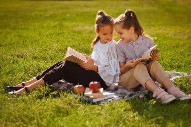 Doğada yeşil çimlerin üzerinde oturmuş kitap okuyan iki genç kız. Arkadaşlar yaz parkında eğleniyorlar.
