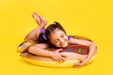 Yaz zamanı çocuk aktivitesi içeriği. Sarı arka planda şişirilebilir yüzüklü mayo giyen küçük kız.
