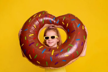 Güneş gözlüklü kız yüzü, büyük kahverengi şişirilebilir halkadan, izole edilmiş donut şeklinde sarı arka plana bakar.