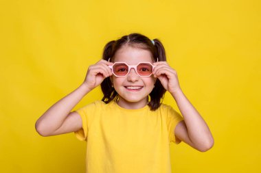 Pembe retro güneş gözlüklü sarı tişörtlü çocuk parlak sarı arka planda kameraya bakıyor. Stüdyo reklam fotoshoot