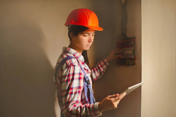 女性の電気技師ビルダーのエンジニアは手のタブレットを握るヒューズ箱の自動スイッチを点検します — ストック写真