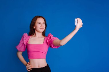 Kabarık kollu pembe elbiseli kız mavi arka planda akıllı telefondan selfie çekiyor.
