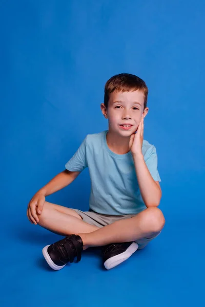 子供の少年は足を横断し ライトブルーのスタジオの背景の上に隔離された青いモックアップTシャツを着用します — ストック写真